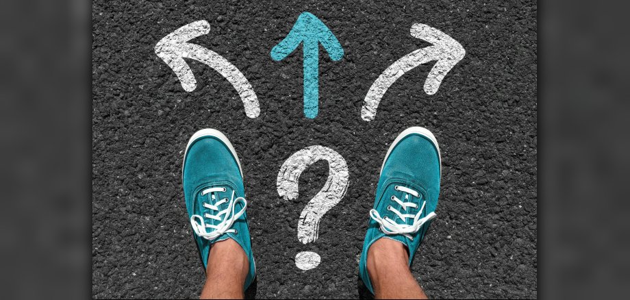 Two men's feet in turquoise trainers on tarmac. In front of him, directional arrows with question marks.