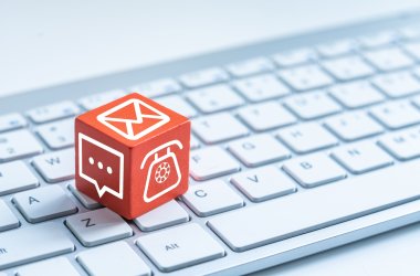 Un cubo rojo con varios símbolos de contacto (teléfono, chat, sobre) descansa sobre un teclado de ordenador blanco.