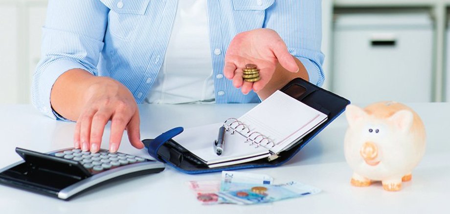 Una mujer lleva dinero en la mano izquierda y maneja una calculadora con la derecha. Delante de ella hay una agenda abierta y un bolígrafo. Sobre la mesa hay varios billetes. A la derecha hay una hucha.
