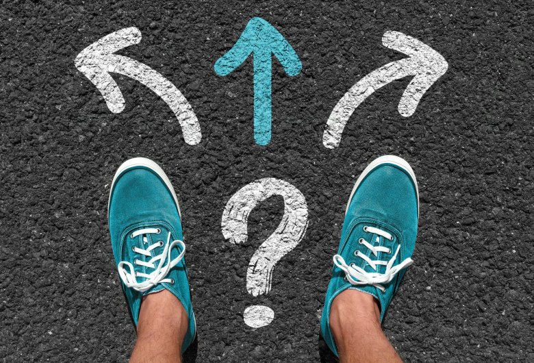 Diferentes formas Dos pies de hombre con zapatillas turquesas sobre el asfalto. Frente a él, flechas de dirección con signos de interrogación.