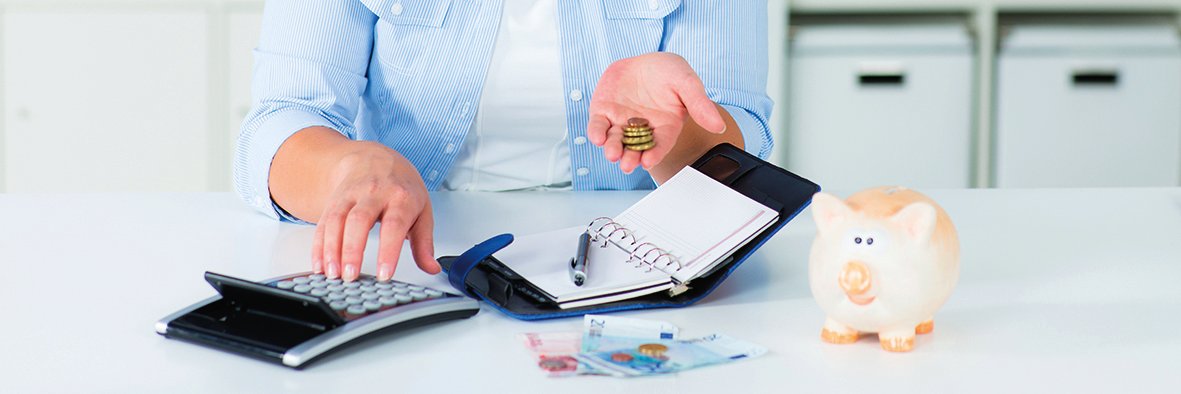Une femme tient de l'argent dans sa main gauche et utilise une calculatrice de la main droite. Devant elle se trouve un organiseur ouvert et un stylo. Plusieurs billets de banque sont posés sur la table. Une tirelire est placée à droite.